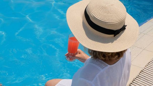 hembra en un sombrero de paja se relaja cerca de la piscina, bebe un cóctel rosa, de cerca. Concepto de humor de verano - Foto, imagen