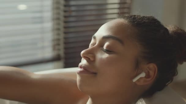 Zblízka portrét klidné uvolněné africké Američanky těší hudbu přes sluchátka, odpočívá ve vaně se zavřenýma očima - Záběry, video