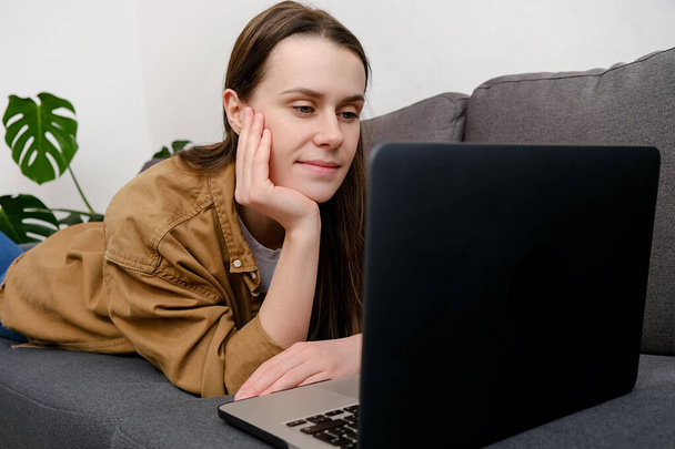 Güzel, beyaz bir kadın evinde rahat bir kanepeye uzanmış dizüstü bilgisayar kullanıyor, internetten arama yapıyor, uzaktan öğreniyor, internette geziniyor ve rahat gri koltukta alışveriş yapıyor. - Fotoğraf, Görsel