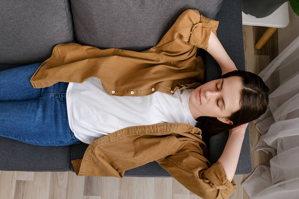 Top view of young woman enjoy day nap on comfy sofa. Caucasian female put hands behind hear lying on cushion on cozy grey couch breath fresh conditioned air inside modern living room. Repose concept - Photo, image