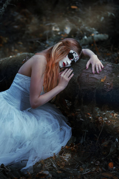 A ghost bride with a frightening make-up on her face in a long white dress sits in a gloomy dark forest. - Foto, imagen