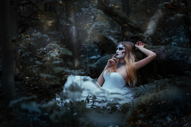 A ghost bride with a frightening make-up on her face in a long white dress sits in a gloomy dark forest. - Foto, afbeelding