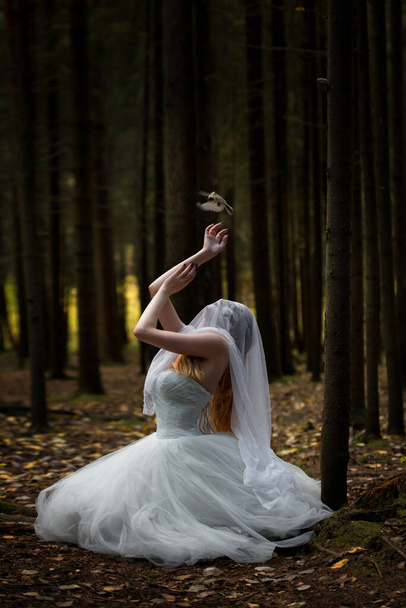 Una misteriosa sposa donna con un volto velato in una cupa foresta in posizione di preghiera con un uccello che striscia nella sua mano. - Foto, immagini