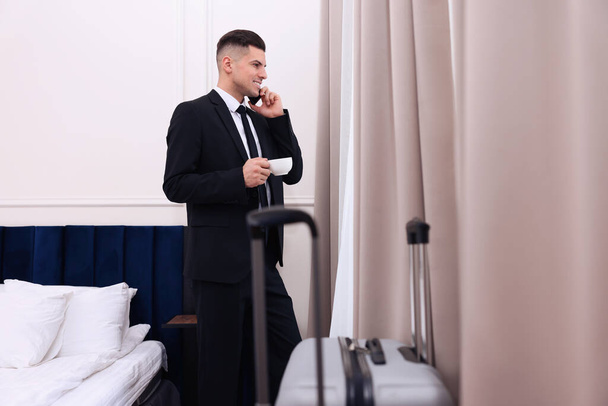 Handsome businessman drinking coffee while talking on phone near window in hotel room - Foto, Bild