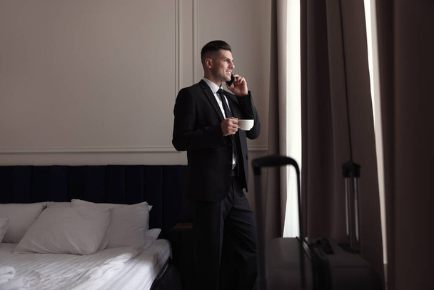 Handsome businessman drinking coffee while talking on phone near window in hotel room - Foto, imagen