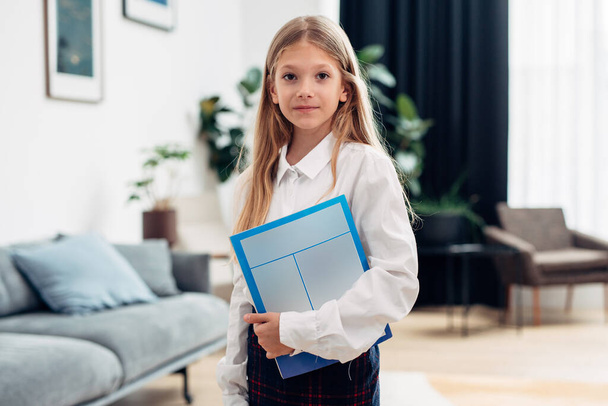 Basisschoolmeisje staat thuis met een leerboek en kijkt naar de camera - Foto, afbeelding