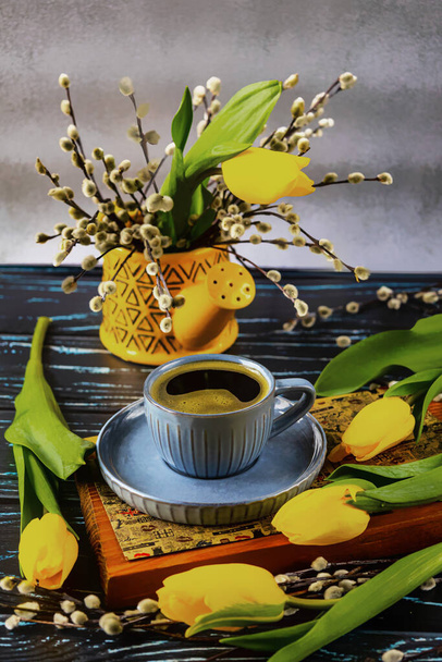 Tazza blu di caffè, tulipani gialli e rami di salice. Annaffiatoio giallo. Natura morta con fiori primaverili. Vista laterale. - Foto, immagini