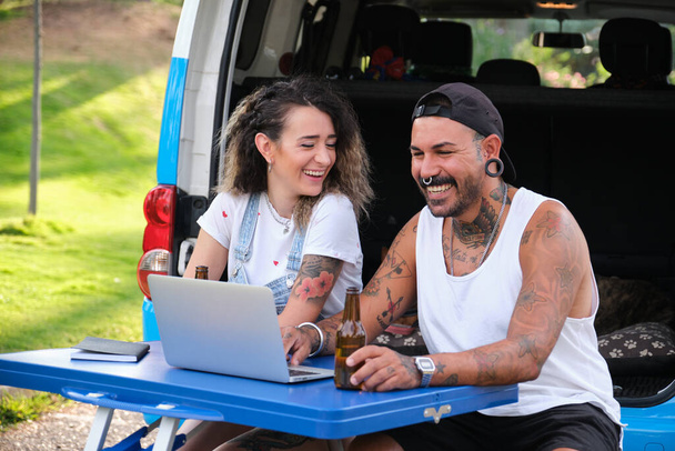 Young tattooed couple laugh and use the laptop in a fold table near by the van. - Photo, Image