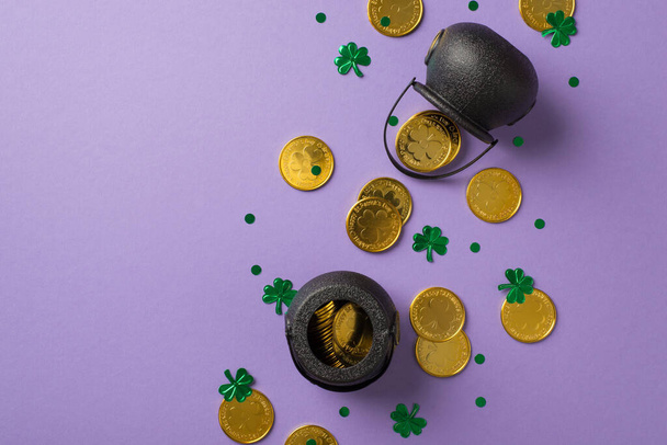 Foto vista superior de dos calderos negros con monedas de oro y confeti en forma de tréboles y puntos sobre fondo vacío violeta aislado - Foto, imagen