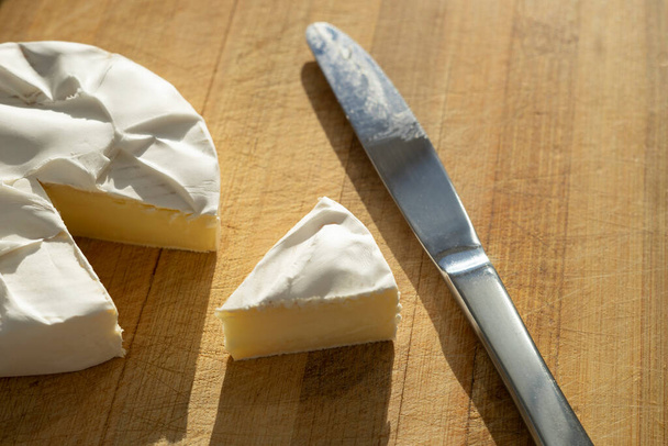Plátky sýra Camembert a celé se stříbrným nožem na slunné dřevěné desce. - Fotografie, Obrázek