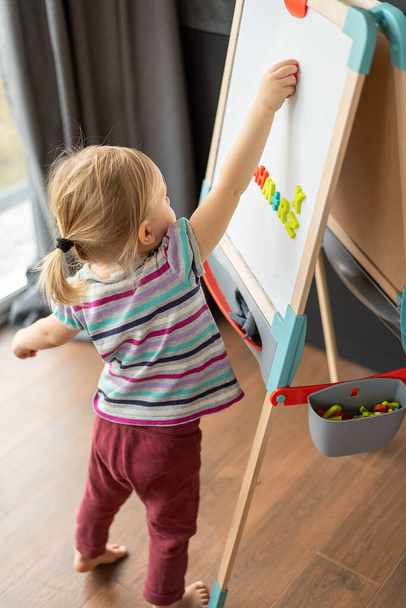 a little fair-haired girl stands near a magnetic board on a childrens easel - Foto, immagini