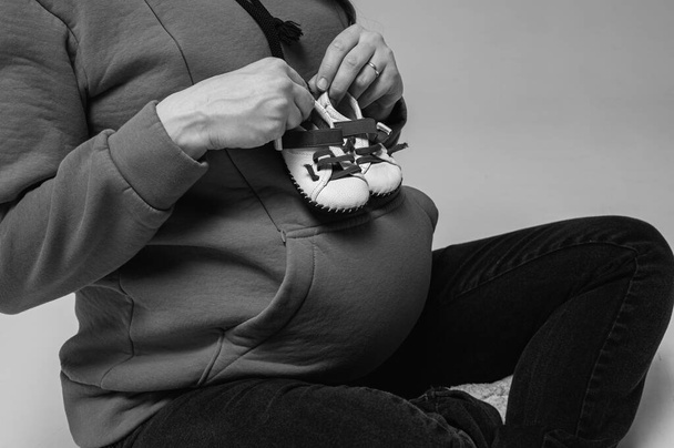 black and white photo, young mother in a hoodie holding small shoes on a big belly, expecting a baby. Isolated on gray background. The gestation period is 38 weeks. Expecting a baby. - Фото, изображение