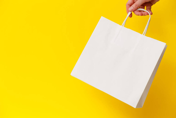 Жіноча рука тримає білу порожню сумку для покупок ізольовано на жовтому тлі. Чорна п'ятниця продаж, знижки, переробка, покупки та екологічна концепція
 - Фото, зображення