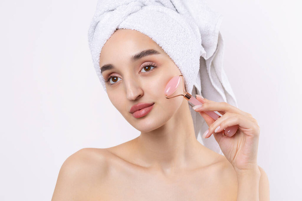 Beautiful girl with thick eyebrows and perfect skin at white background, towel on head, beauty photo. Holds a cosmetic roller. - Photo, Image