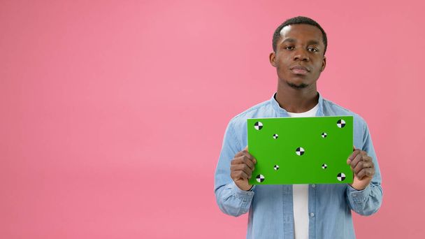 Serious African-American holding Banner with Green Screen Tracks Points for Copy Space. Empty Green Screen Board. Does Not Smile Looks at Camera With Space for Text or Advertising on Pink Background. - Фото, зображення