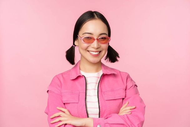 Stylish asian girl in pink clothes and sunglasses, smiling and looking happy at camera, standing over studio background - Zdjęcie, obraz