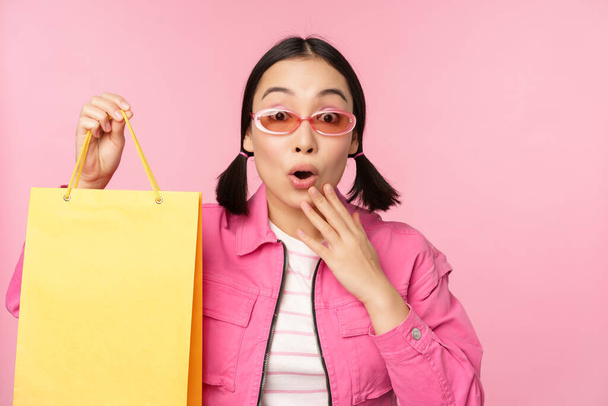 Nakupovat. Stylová asijská dívka v slunečních brýlích, ukazuje tašku z obchodu a usmívá se, doporučuje prodej promo v obchodě, stojící nad růžovým pozadím - Fotografie, Obrázek