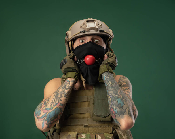 een soldaat in militaire kleding helm met een bdsm gag in zijn mond drukt emoties, foto grap - Foto, afbeelding