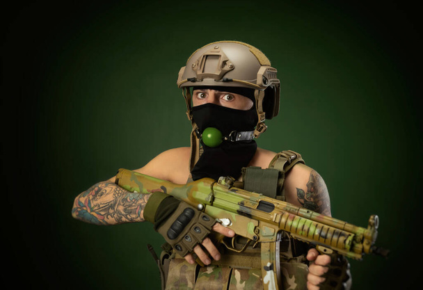 un soldato di sesso maschile in abiti militari, un casco con un bdsm bavaglio in bocca esprime emozioni tenendo in mano un fucile automatico, foto scherzo - Foto, immagini