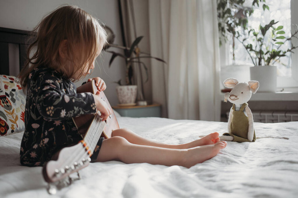 toddler girl playing ukulele guitar sitting on the bed at home - Φωτογραφία, εικόνα