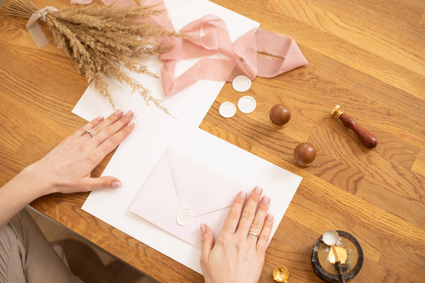 Przycięte zdjęcie kobiecych rąk, dzięki czemu chwasty koperty zaproszenia rzemiosła, ozdobić różową wstążką i woskiem fok. Widok z góry - Zdjęcie, obraz