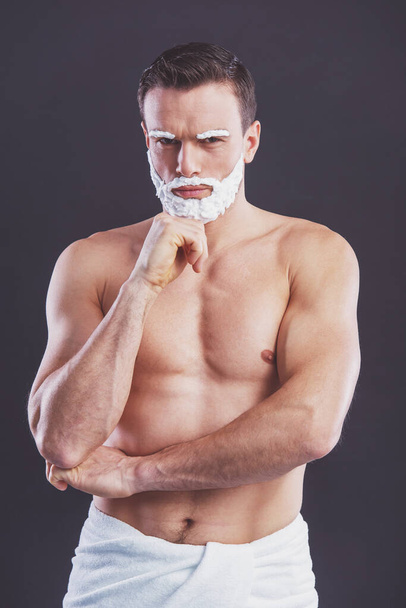 Handsome naked man with shaving foam on his face is looking at camera, standing with his hand on chin on a dark background - Foto, Imagem