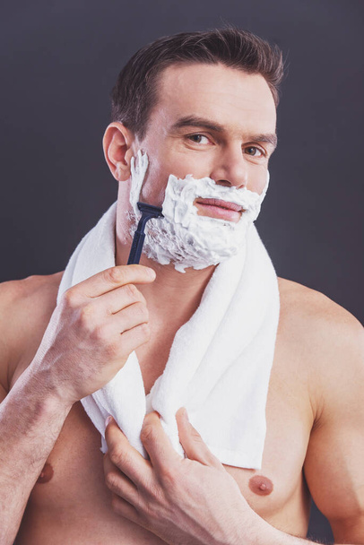 Portrait of handsome naked man with a towel on his shoulders shaving using a razor and looking at camera, on a dark background - Photo, image