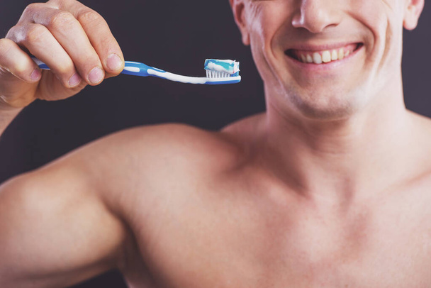 Portrait of handsome naked man holding a toothbrush with toothpaste and smiling, on a dark background, close up - Zdjęcie, obraz