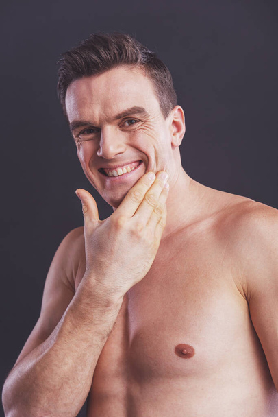 Portrait of handsome naked man touching his shaved face, looking at camera and smiling, on a dark background - Foto, afbeelding