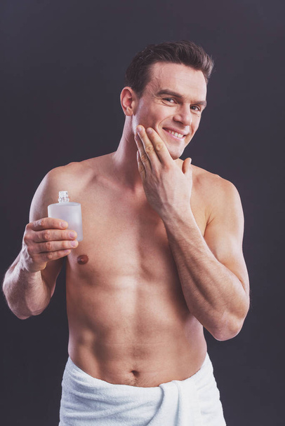 Handsome naked man is smiling, holding aftershave lotion and applying it, on a dark background - Foto, Imagen