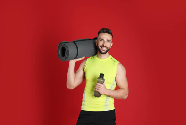Handsome man with yoga mat and bottle of water on red background - Photo, Image