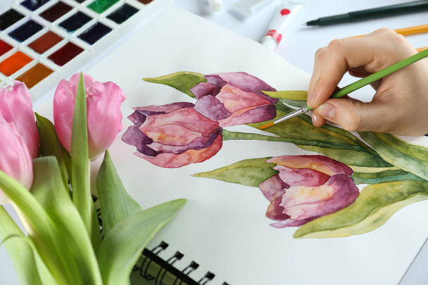 Woman painting tulips in sketchbook at white table, closeup - Photo, image