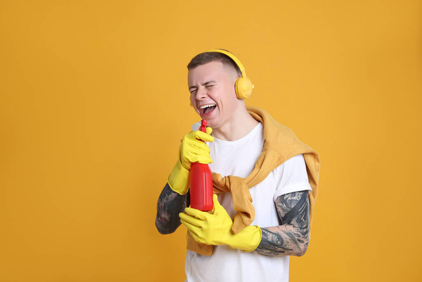 Handsome young man with bottle of detergent singing on orange background. Space for text - 写真・画像