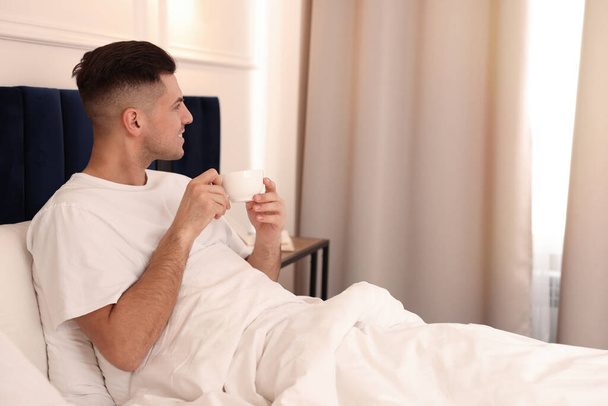 Красивый мужчина пьет кофе на кровати в гостиничном номере - Фото, изображение