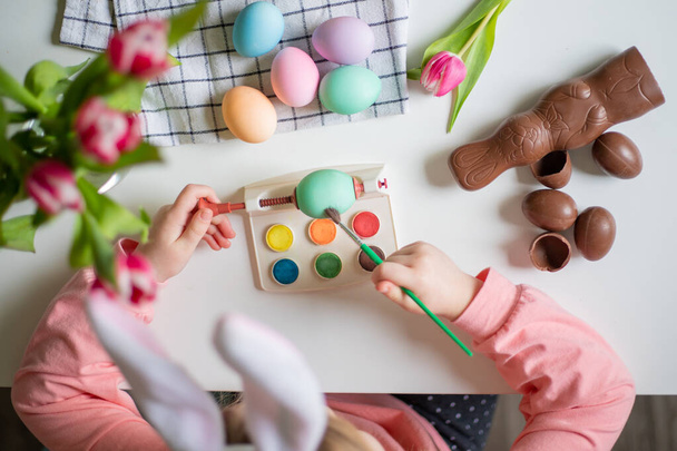 Top kilátás egy kislány nyuszi fülek gazdaság ecset és díszítő húsvéti tojás akvarell. - Fotó, kép