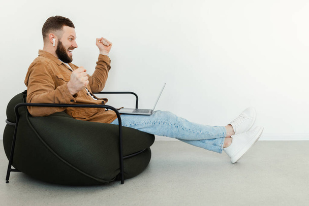 Emocjonalny człowiek za pomocą laptopa wstrząsające pięści siedzi w fotelu w pomieszczeniach - Zdjęcie, obraz