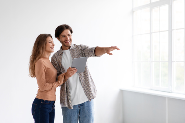 Cheerful millennial european male and female planning new interior with tablet in empty room of new apartment - Foto, immagini