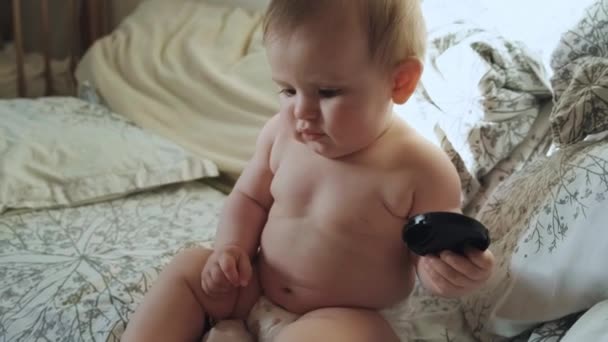 赤ん坊はベッドの上に彼の足でぬいぐるみのおもちゃを押す遊び心のあるおむつだけで裸に座っている。美肌ケア。赤ん坊の世話だ。美顔だ。家族の日だ。幸せな顔. - 映像、動画