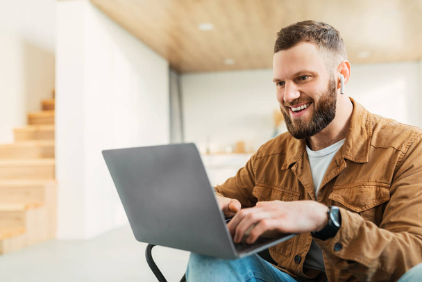 masculino animado usando laptop usando fones de ouvido trabalhando on-line em casa - Foto, Imagem