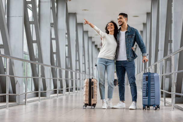 Reisewerbung. Glückliches Paar aus dem Nahen Osten steht auf Flughafen und zeigt daneben - Foto, Bild