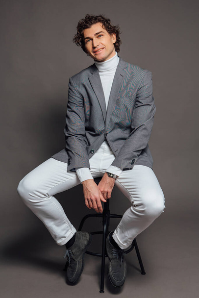 Fashion portrait of a tall handsome man dressed in white turtleneck, white jeans and grey jacket sitting on chair and posing on the grey background - Foto, Imagen