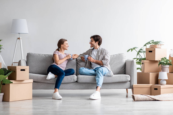 Frohes junges europäisches Paar entspannt auf dem Sofa, trinkt Kaffee, redet und plant neues Interieur im Zimmer - Foto, Bild
