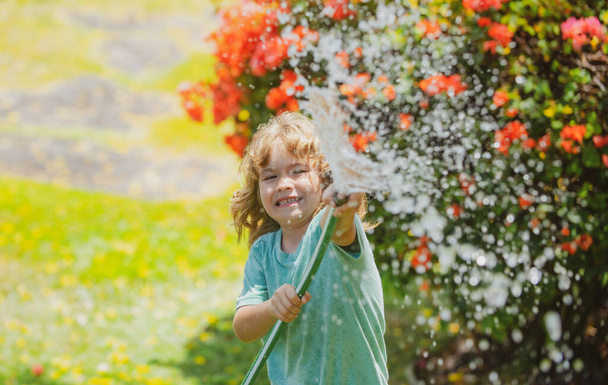 Cute little boy watering flowers in the garden at summer day. Child using garden hose. Funny kid watering plants in the yard garden. - Фото, изображение