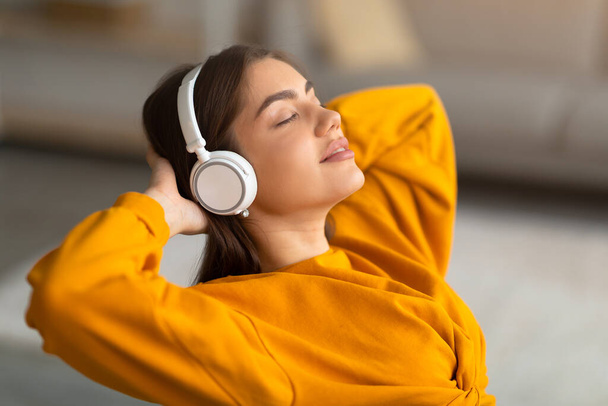 Spokojna młoda kaukaska kobieta słuchająca muzyki w słuchawkach bezprzewodowych, relaksująca się z rękami za głową w domu - Zdjęcie, obraz