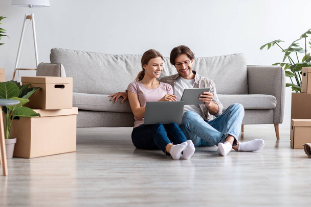Glad smiling caucasian husband and wife in casual planning new interior with tablet and computer in living room - Photo, Image