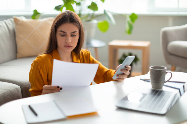 Porträt einer gestressten jungen Frau, die online arbeitet, Finanzdokumente liest, Laptop und Handy zu Hause benutzt - Foto, Bild