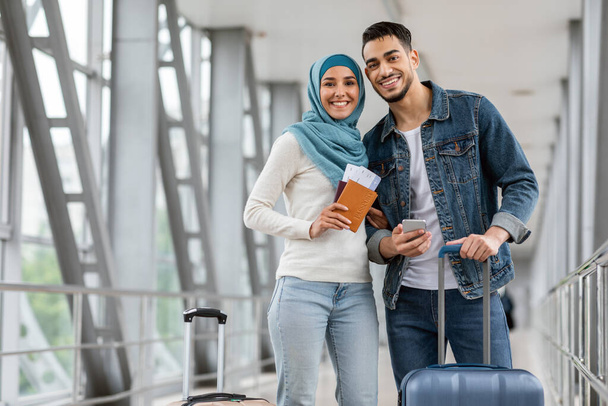 Retrato de feliz pareja islámica de pie en el aeropuerto con equipaje y teléfono inteligente - Foto, Imagen