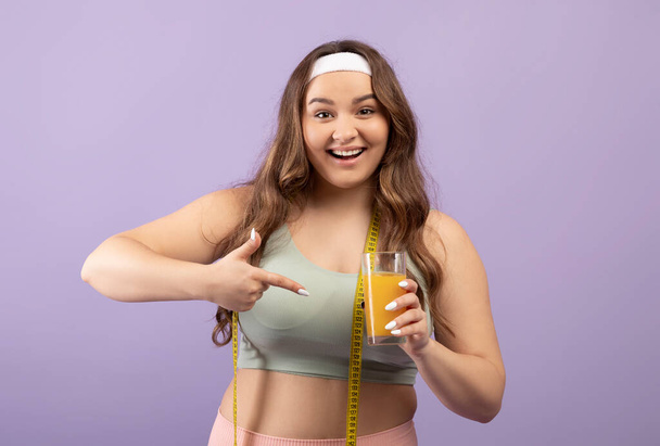 Glad smiling plus size european millennial lady in sportswear with measuring tape points finger at glass of orange juice - Fotografie, Obrázek