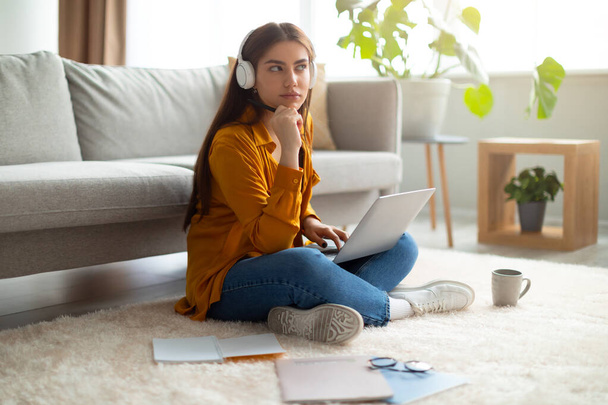 Videollamada en línea. Mujer joven reflexiva en auriculares sentados en el suelo, utilizando el ordenador portátil, teniendo conferencia virtual en interiores - Foto, Imagen