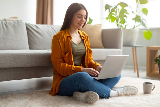 Piena lunghezza di felice giovane donna seduta gambe incrociate sul pavimento con computer portatile, lavorare o studiare online a casa - Foto, immagini